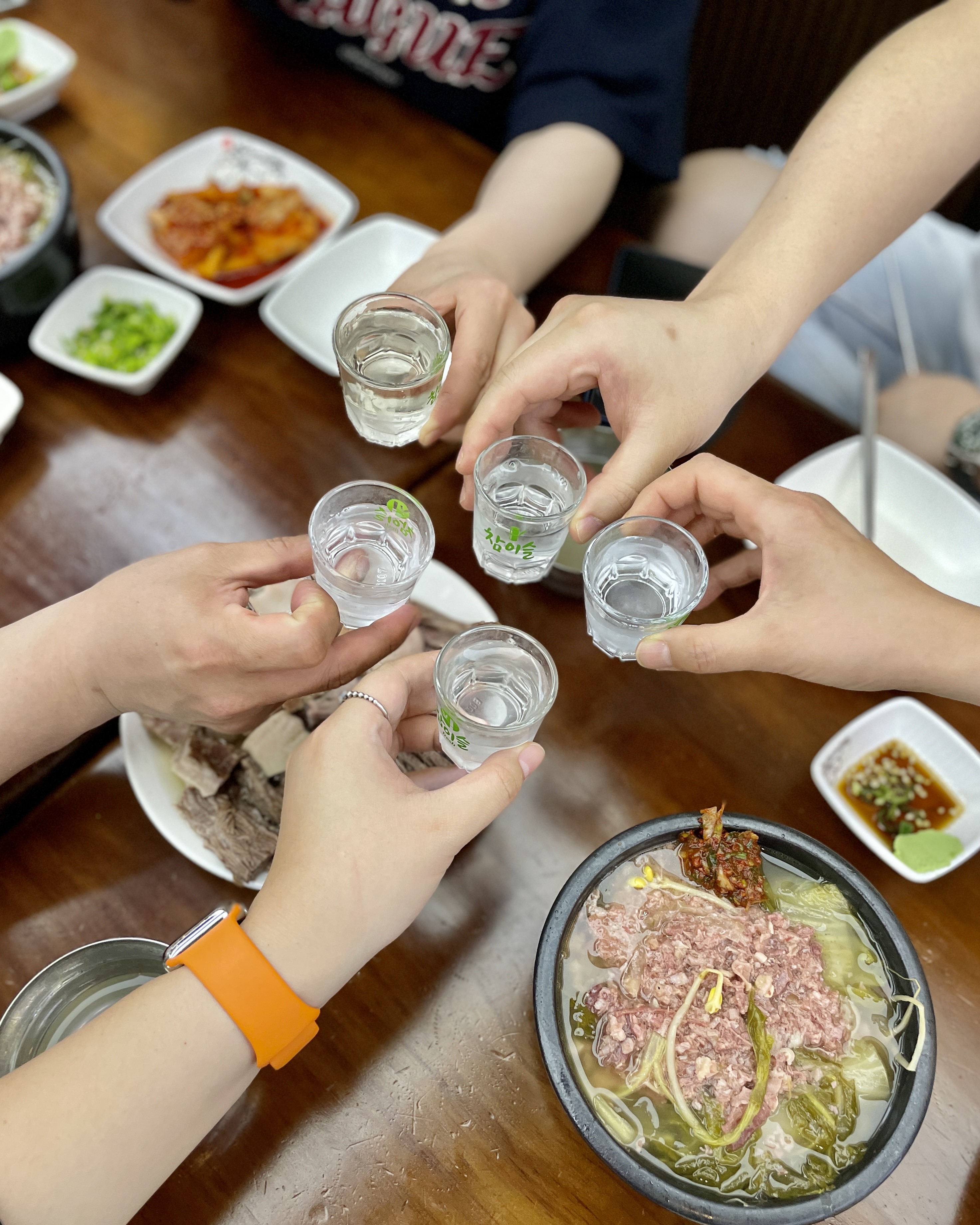 해장국과 수육이 놓인 식탁 위로 여러 손이 잔을 모은 장면